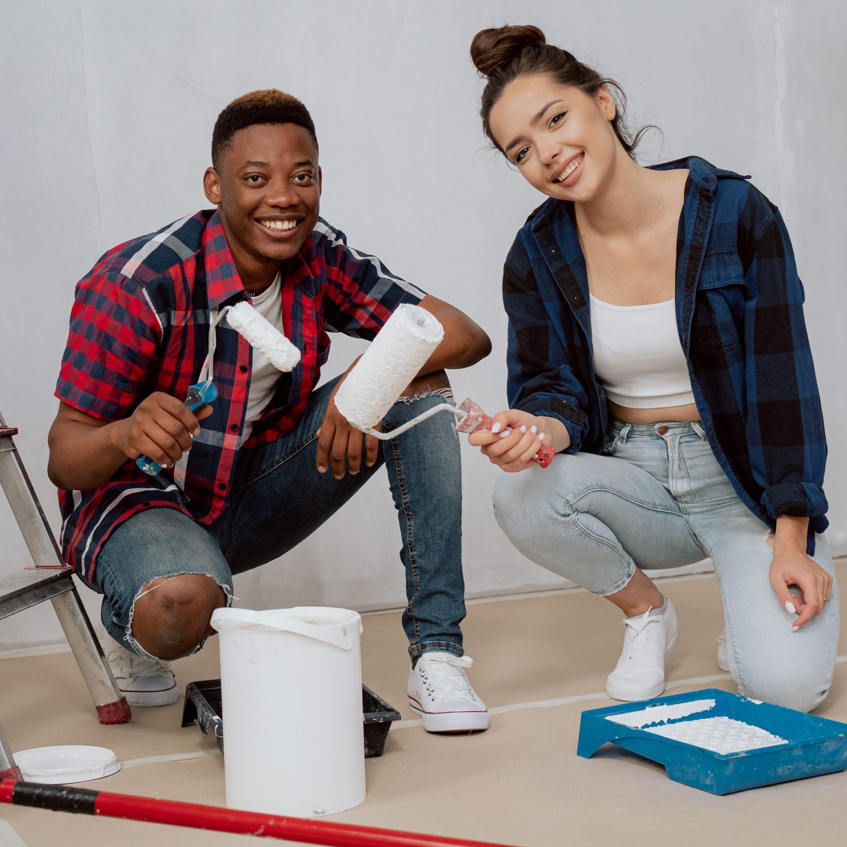 Two happy young people in love are renovating their first purchased apartment, they are kneeling on floor sealed with cardboard scooping paint from paint tray they are holding rollers in hands